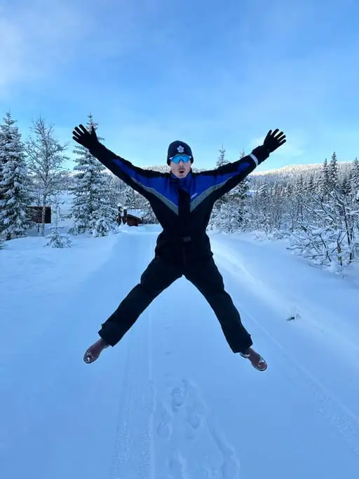 A man in a black and blue snowsuit mid-star-jump on a snow-covered track, arms and legs fully extended, snow-laden pines behind him.