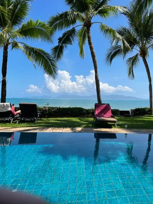 An infinity pool looking out past palm trees to a calm sea and a distant island under white cloud.