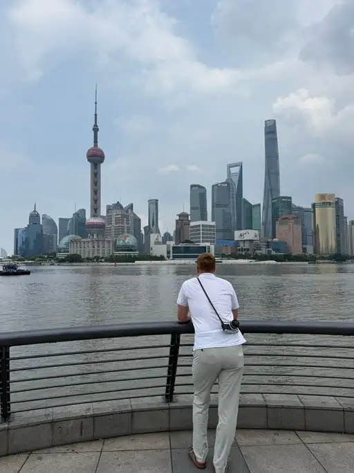 A man in a white t-shirt leaning on a riverside railing, looking across the water at the Pudong skyline and the Oriental Pearl Tower.
